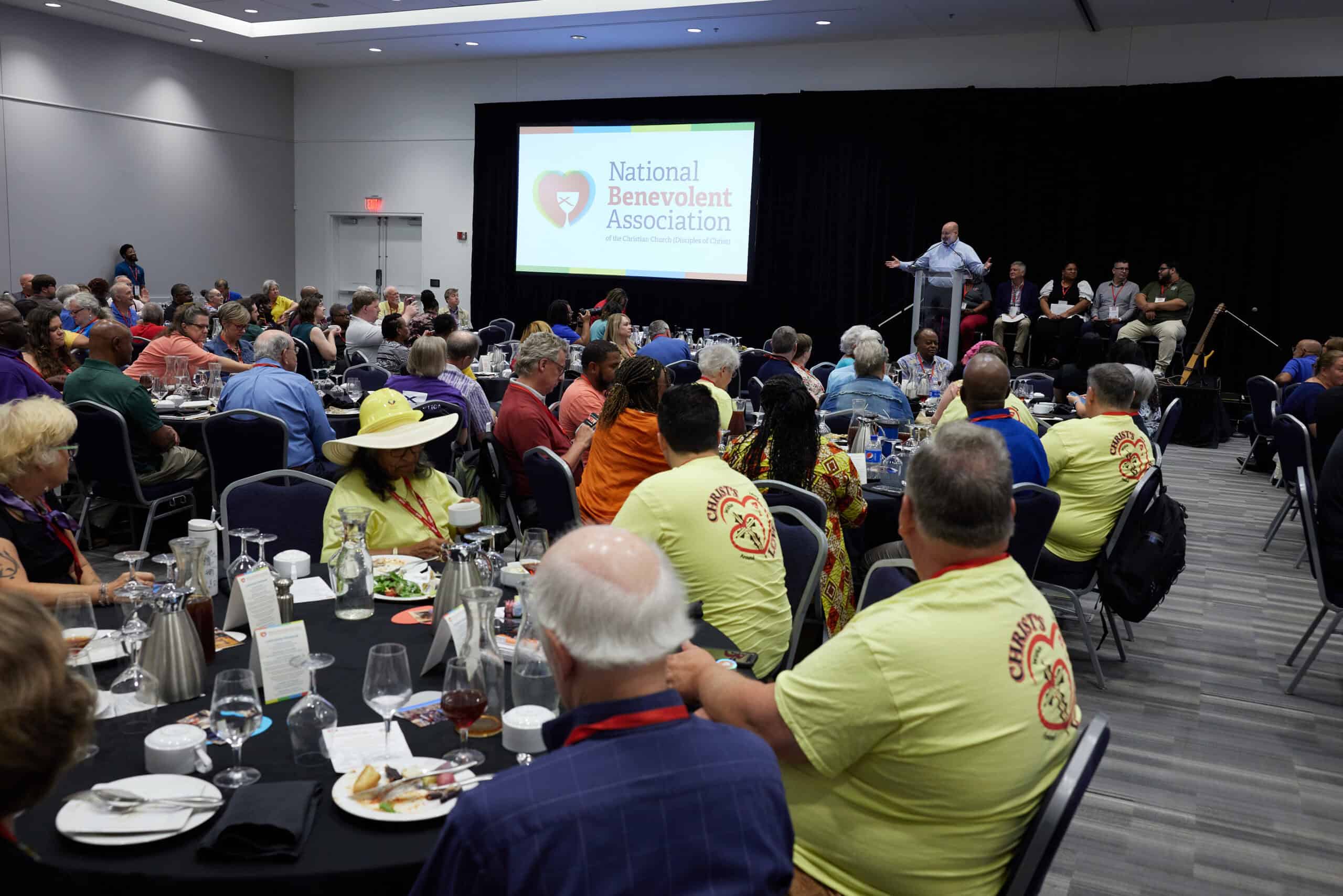 Man speaking onstage to an audience seated at tables inside a conference room.